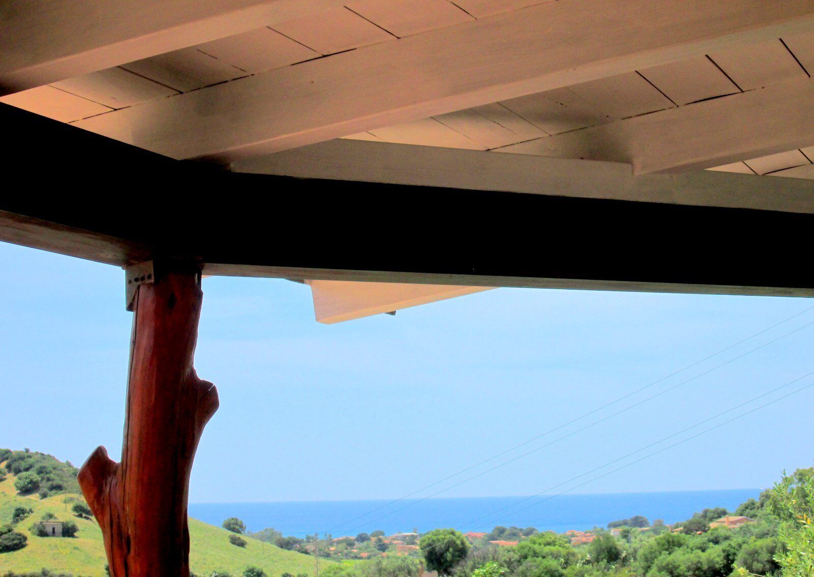 Blick durch eine Holzhütte mit geneigtem Dach auf eine Küstenlandschaft mit Hügeln und dem Meer.