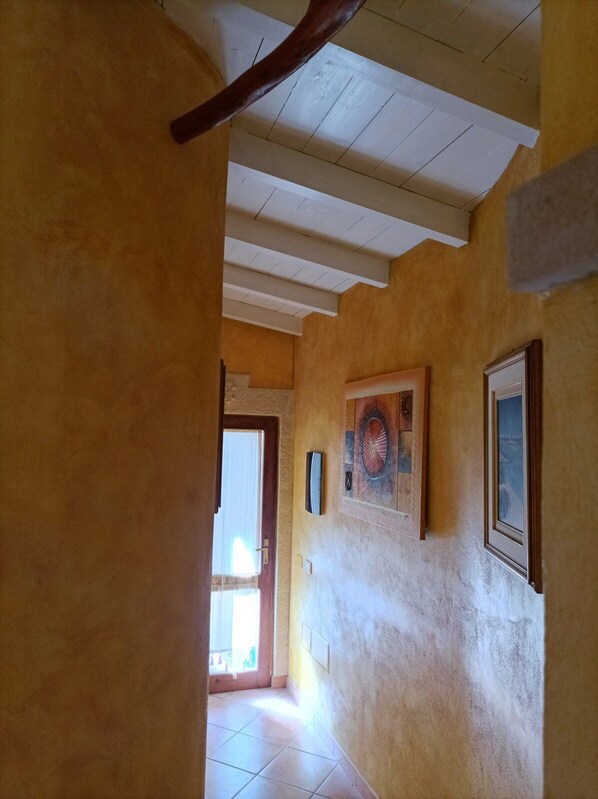 A hallway with a wooden ceiling, a door leading to a bright room, and a wall with framed artwork.