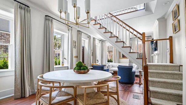 Un amplio salón con una mesa redonda, sillas de mimbre, un sofá azul y una escalera con barandilla de madera.