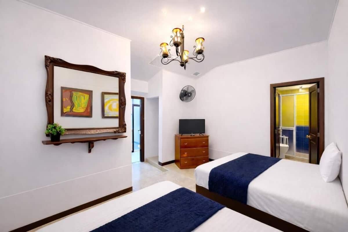 Ein Hotelzimmer mit zwei Betten, einem Fernseher, einer Chandelüster, einer Uhr und Bildern an der Wand.