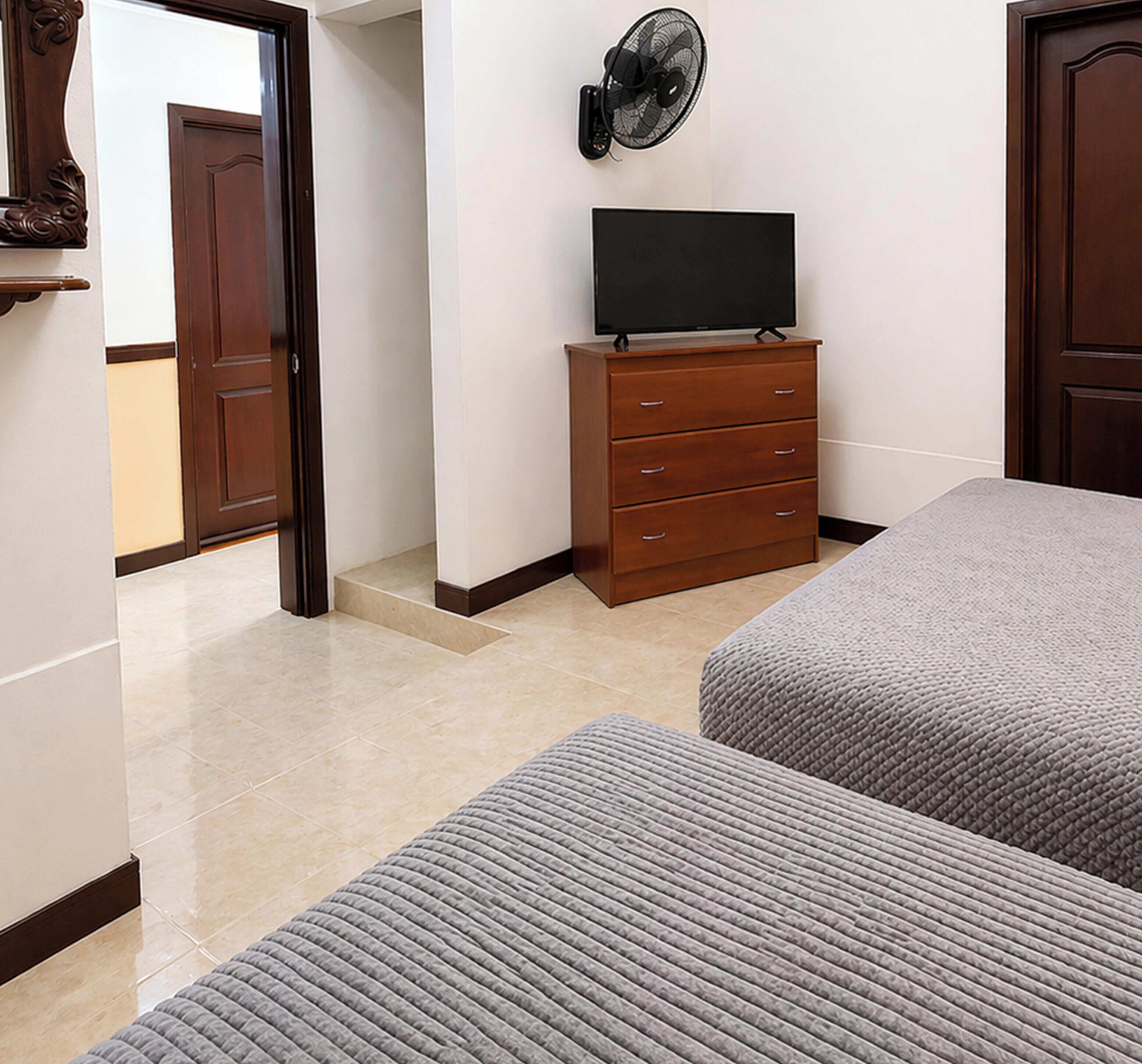 Un dormitorio con una televisión sobre una cómoda, dos camas, un ventilador y una puerta que da a otra habitación.