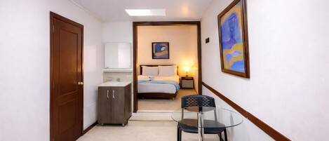 A narrow hallway leading to a bedroom with a bed, bedside table, and a nightstand. There is a small table with chairs in the hallway.