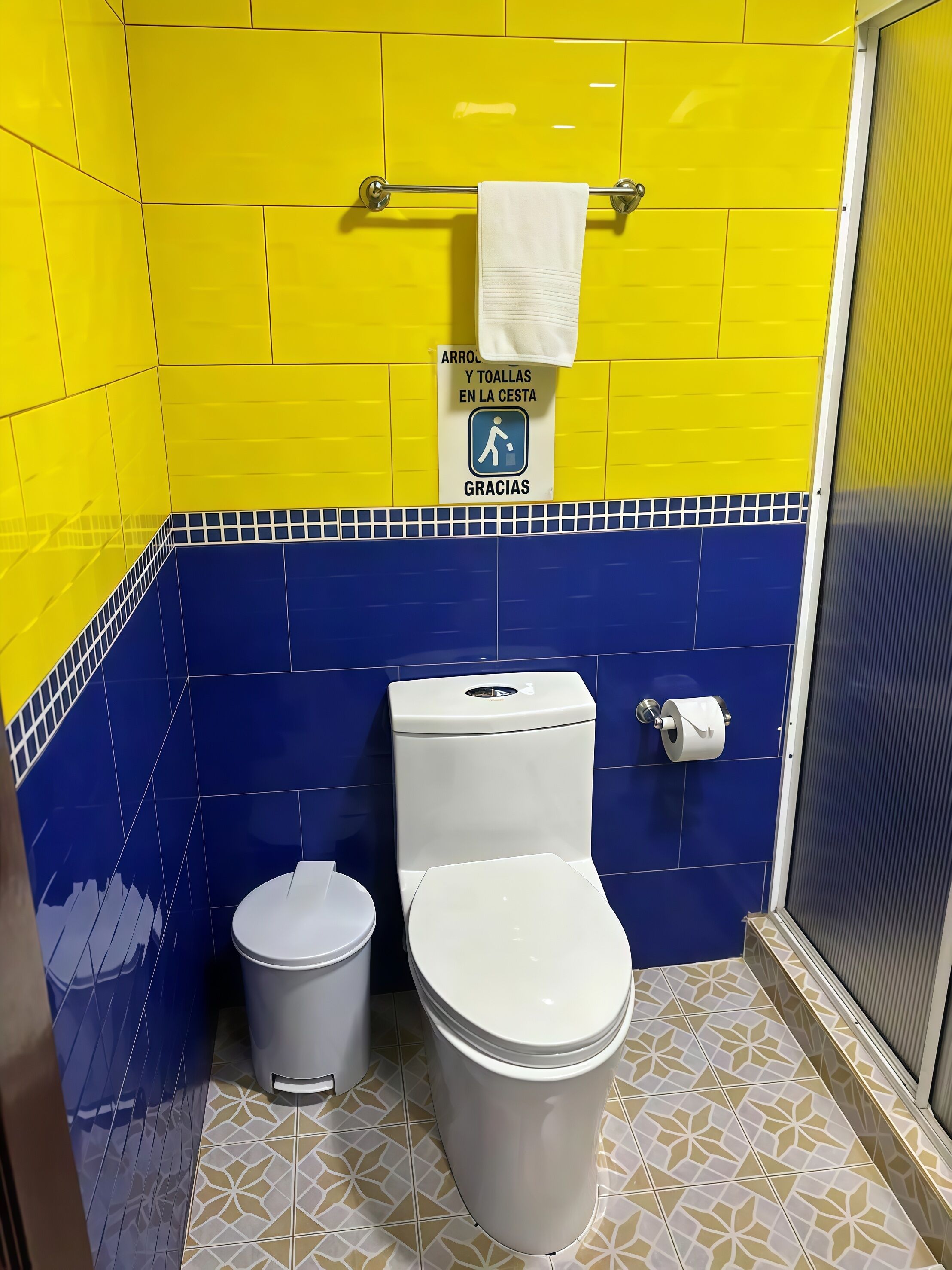 A bathroom with yellow and blue tiles, a toilet, a trash bin, and a hand dryer.
