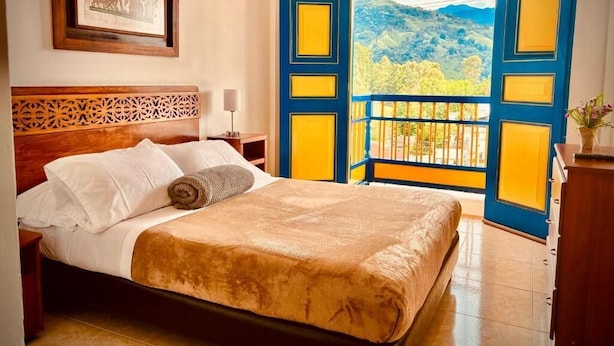 A bed with white linens and a brown blanket, a wooden headboard, a framed picture on the wall, and a balcony with blue and yellow doors. - Hospedaje Camino Real  (Salento)