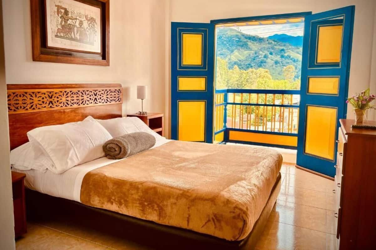 Ein Bett mit weißen Laken und einer braunen Decke, ein hölzernes Kopfteil, ein gerahmtes Bild an der Wand und ein Balkon mit blauen und gelben Türen.