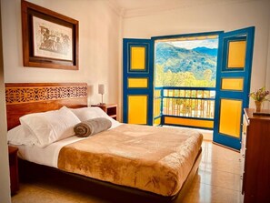 Ein Bett mit weißen Laken und einer braunen Decke, ein hölzernes Kopfteil, ein gerahmtes Bild an der Wand und ein Balkon mit blauen und gelben Türen.