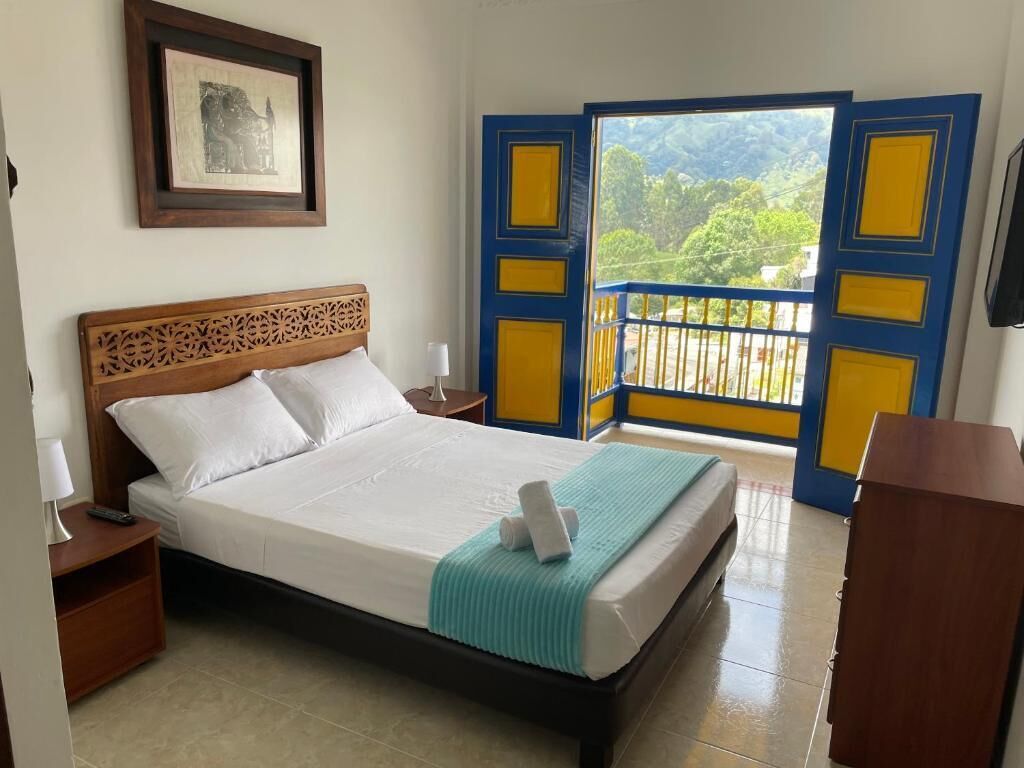 Habitación de hotel con cama, mesita de noche, cuadro en la pared y balcón con vistas a zonas verdes.