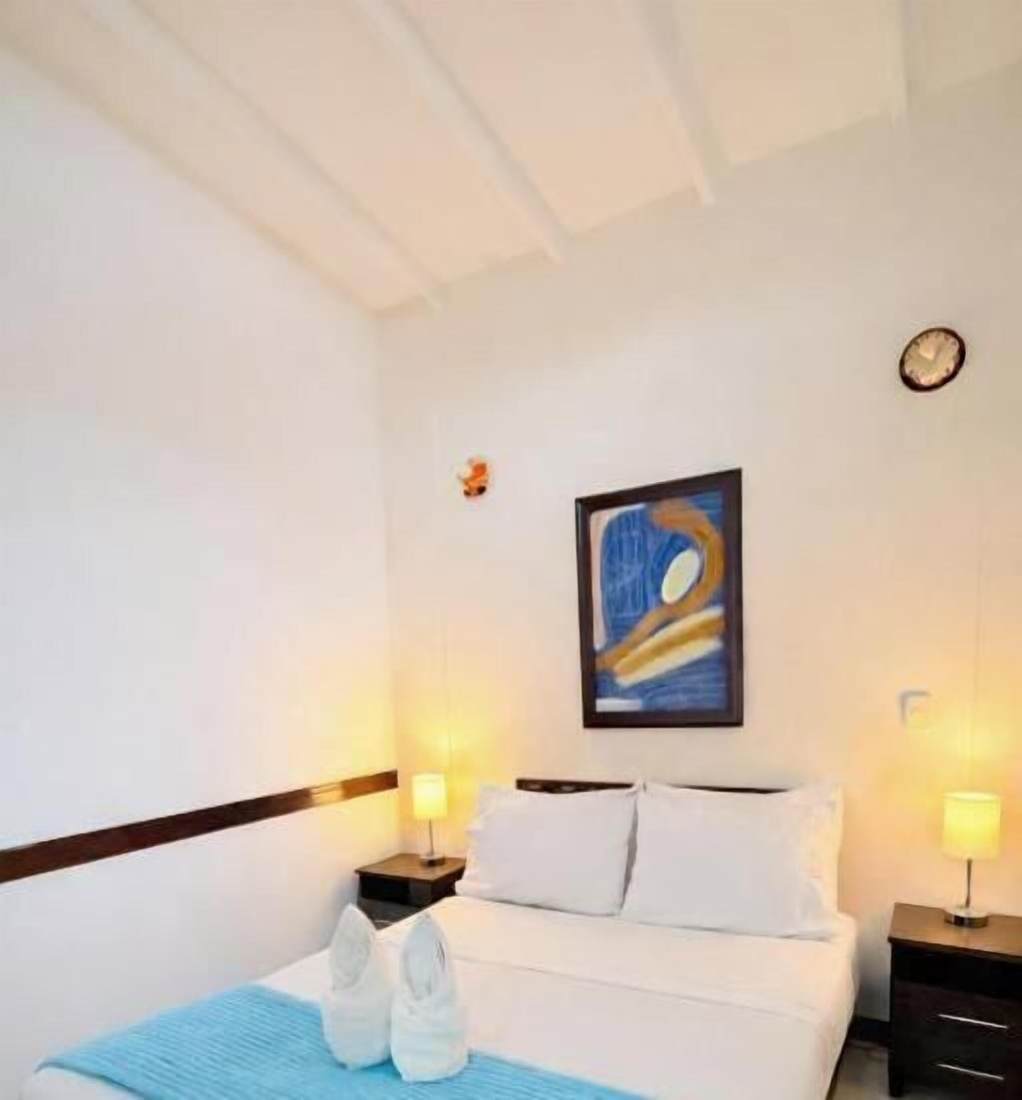 Habitació d'hotel amb un llit, dues taules de nit amb làmpades, un quadre a la paret i un rellotge.