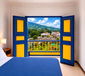 A hotel room with a bed, a nightstand, a lamp, and a balcony with a view of a hillside town. - Hospedaje Camino Real  (Salento)