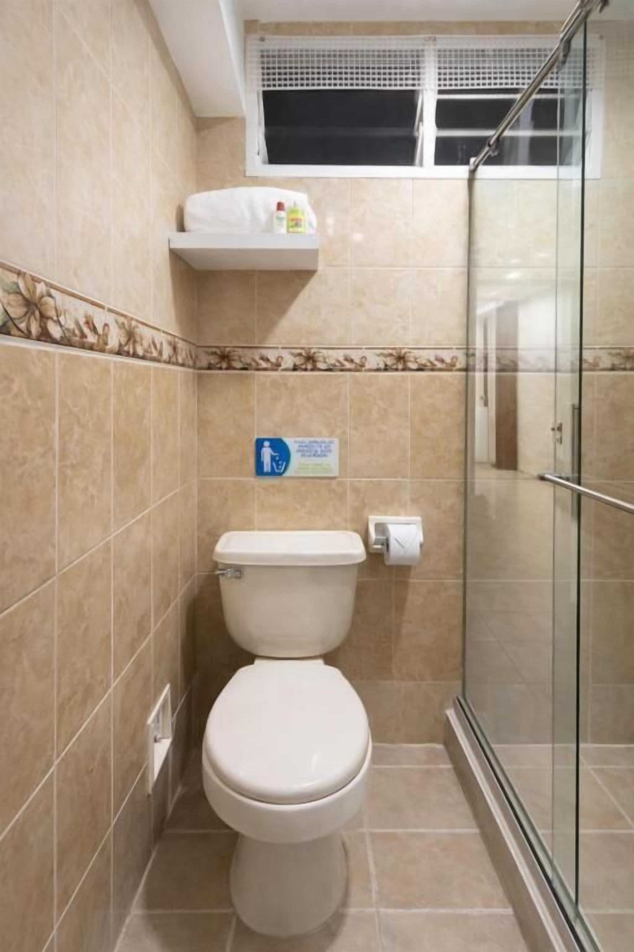 Une salle de bain équipée de toilettes, d’une douche à parois de verre et d’un distributeur de gel hydroalcoolique placé sur une étagère.
