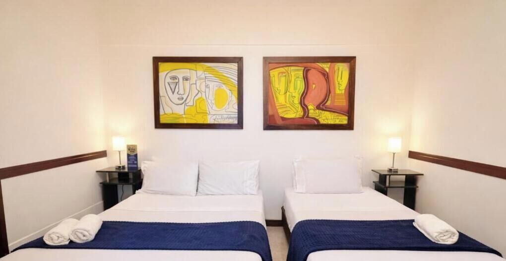 A hotel room with two beds, white bedding, blue throw blankets, and artwork on the walls.