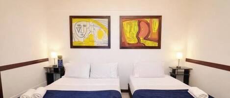 Habitación de hotel con dos camas, ropa de cama blanca, mantas azules y cuadros en las paredes.