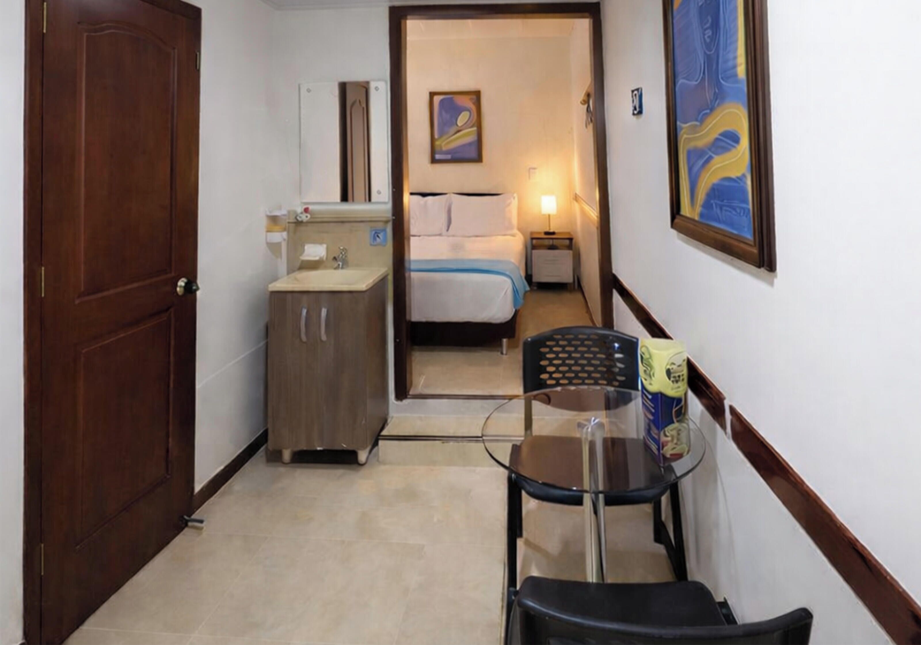 A hotel room with a wooden door, a small kitchenette, a bed with white linens, a nightstand with a lamp, and a painting on the wall.