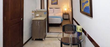 A hotel room with a wooden door, a small kitchenette, a bed with white linens, a nightstand with a lamp, and a painting on the wall.