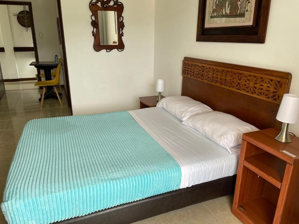 A neatly made bed with a blue and white bedspread, a wooden headboard, a bedside table with a lamp, and a framed picture on the wall.
