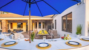 An outdoor dining area with a round table set for six, under a blue umbrella. - Epic Resort: Pool, Hot Tub, Pickleball & More! (Chandler)