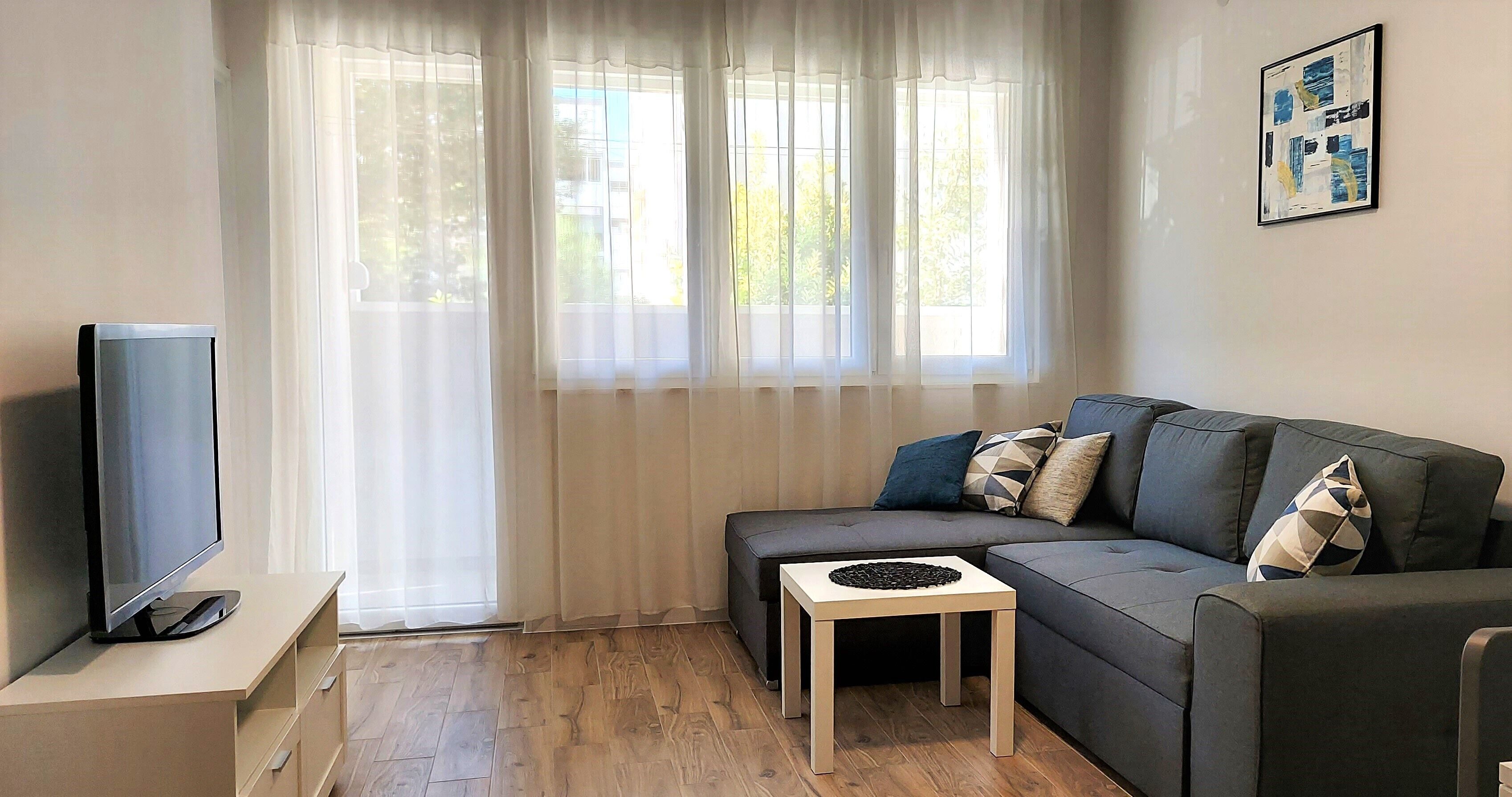A modern living room with a flat-screen TV, a grey sofa with patterned cushions, a small wooden side table, and a large window with sheer curtains.
