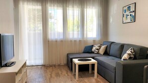 A modern living room with a flat-screen TV, a grey sofa with patterned cushions, a small wooden side table, and a large window with sheer curtains. - Apartment Adora Split (Split)
