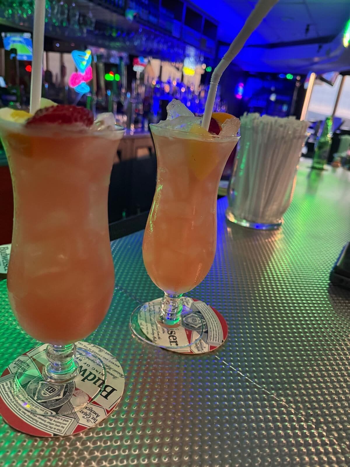 Two cocktails on a bar counter with a textured surface and coaster with text.