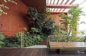 A wooden deck with a metal railing, surrounded by lush greenery and a red wall.