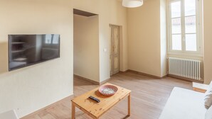 A modern living room with a flat-screen TV, a wooden coffee table with a bowl and remote, a white sofa, and a large window with a view. - PM7        (Castello Cabiaglio)