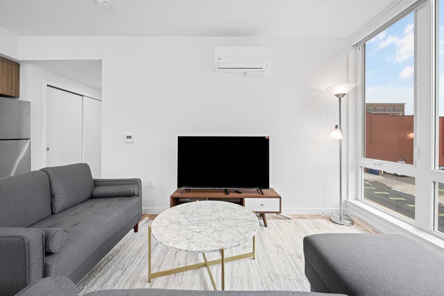 Een moderne woonkamer met een grijze bank, een ronde marmeren salontafel, een flatscreen televisie op een houten dressoir en een groot raam met uitzicht.