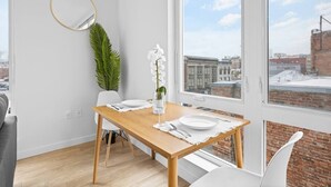 Un comedor con una mesa de madera, sillas blancas, una orquídea blanca en un florero de vidrio y vistas de la ciudad a través de un amplio ventanal.