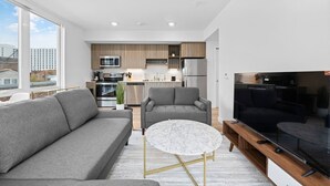 Un apartamento moderno con sala de estar, sofá gris, mesa de centro redonda, televisor y área de cocina con electrodomésticos.