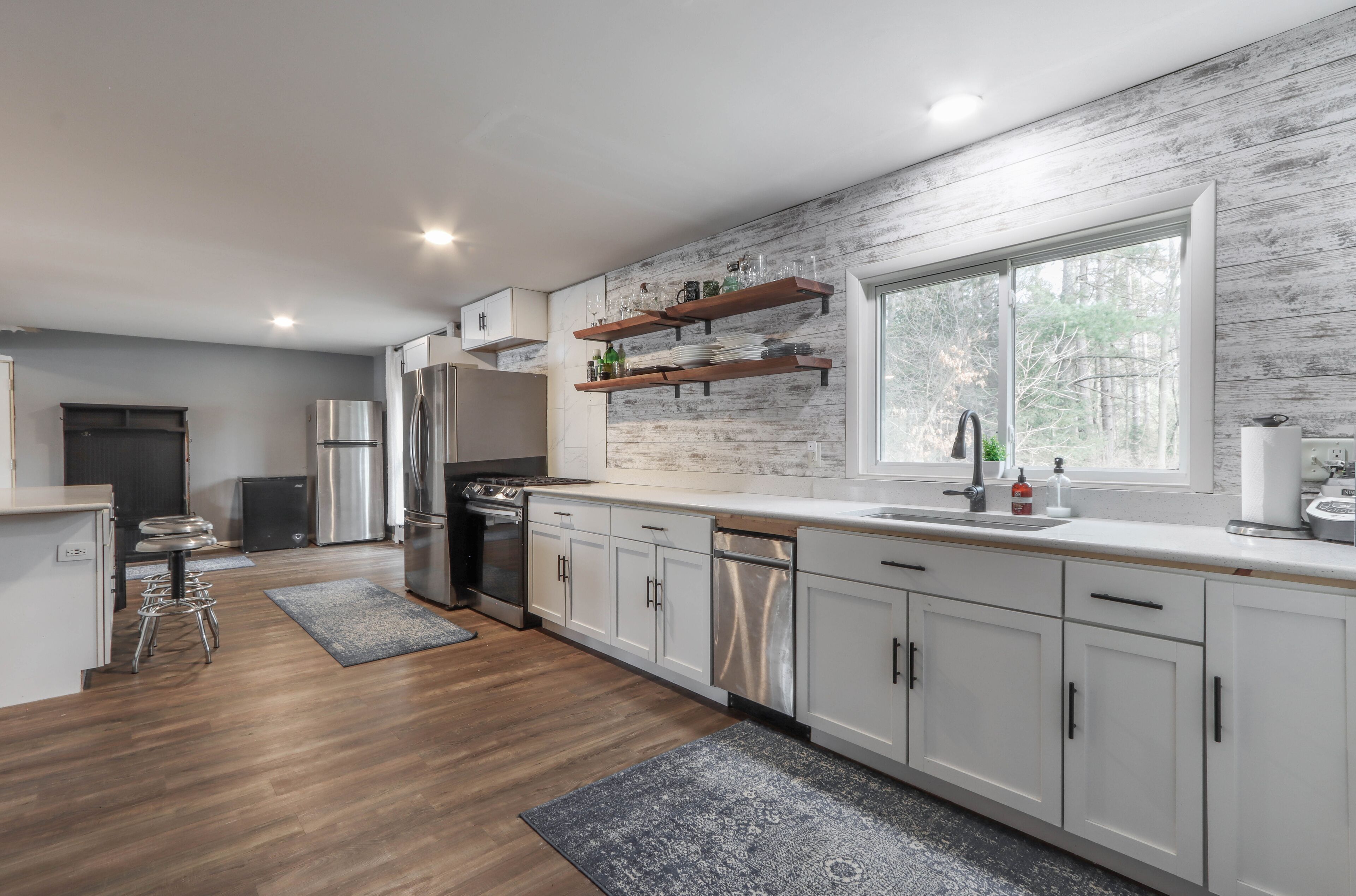 Een moderne keuken met witte kasten, roestvrijstalen apparatuur en een houten vloer. Er staan barkrukken en er is een raam dat voor natuurlijk licht zorgt.