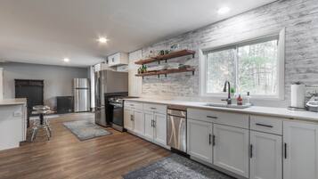 Een moderne keuken met witte kasten, roestvrijstalen apparatuur en een houten vloer. Er staan barkrukken en er is een raam dat voor natuurlijk licht zorgt.