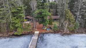 Ein Haus, umgeben von dichtem Wald, eine Anlegestelle, die sich in ein gefrorenes Gewässer erstreckt, und ein kleines Boot an der Anlegestelle.