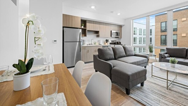 Un apartament modern amb zona de menjador, sofà gris, cuina i grans finestres.