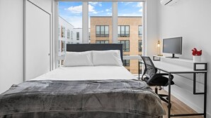 Una habitació d'hotel moderna amb un llit gran, un escriptori amb un ordinador i vista a un paisatge urbà.