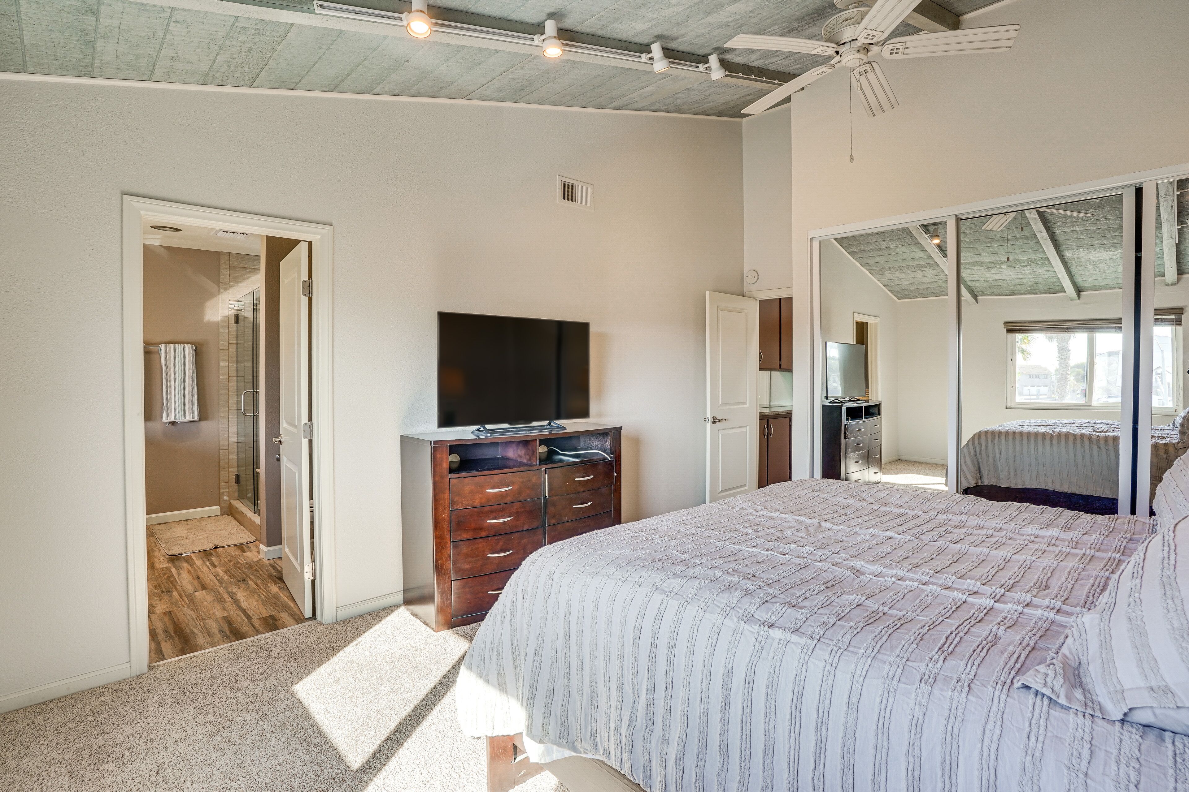 A bedroom with a large bed, a TV on a wooden dresser, a walk-in closet, and a sliding glass door leading to a balcony.
