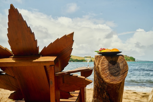 A wooden beach chair and table with a plate of fruit and a glass of wine on a wooden stump, overlooking the sea.
