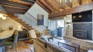 A spacious living area with a stone fireplace, comfortable seating, a dining area, and a staircase with exposed wooden beams. - Mountain-View Condo in Brian Head w/ Private Sauna (Brian Head)