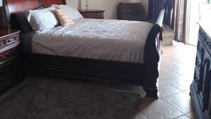 Een slaapkamer met een bed, nachtkastje, dressoir, plafondventilator, spiegel en een raam met gordijnen.