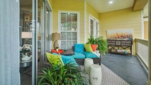 A covered porch with a wicker sofa, colorful cushions, a lamp, and potted plants.