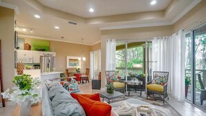 A spacious living area with a sofa, coffee table, and a dining table with chairs. There is a kitchen area with cabinets and a refrigerator. The room has large windows and a sliding glass door leading to a balcony.