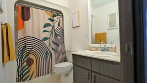 Een badkamer met douchegordijn met bladmotief, toilet, wastafel met spiegel erboven en een handdoekrek met een gele handdoek.