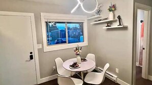 Een eetgedeelte met een ronde tafel en vier witte stoelen, een raam met uitzicht op een parkeerplaats, en een plafondlamp.
