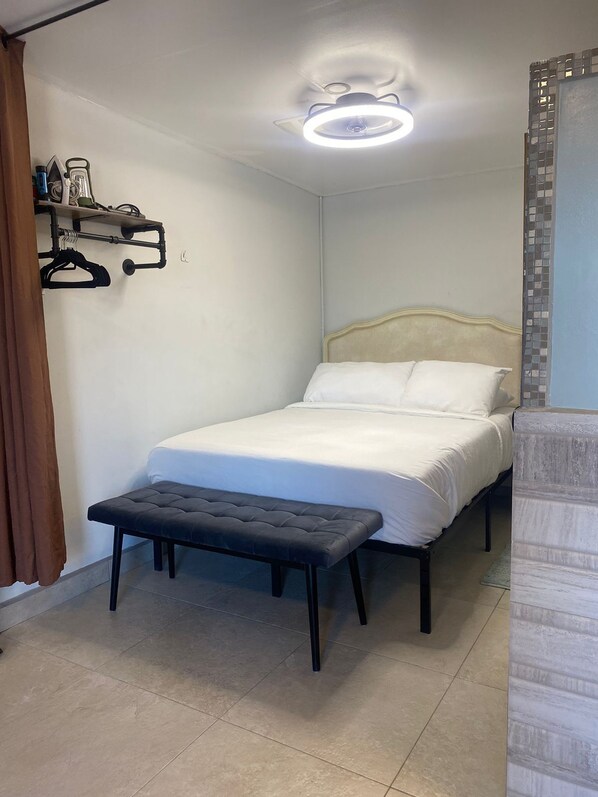 Une chambre à coucher comprenant un lit, un banc, une étagère avec des cintres et un luminaire au plafond.