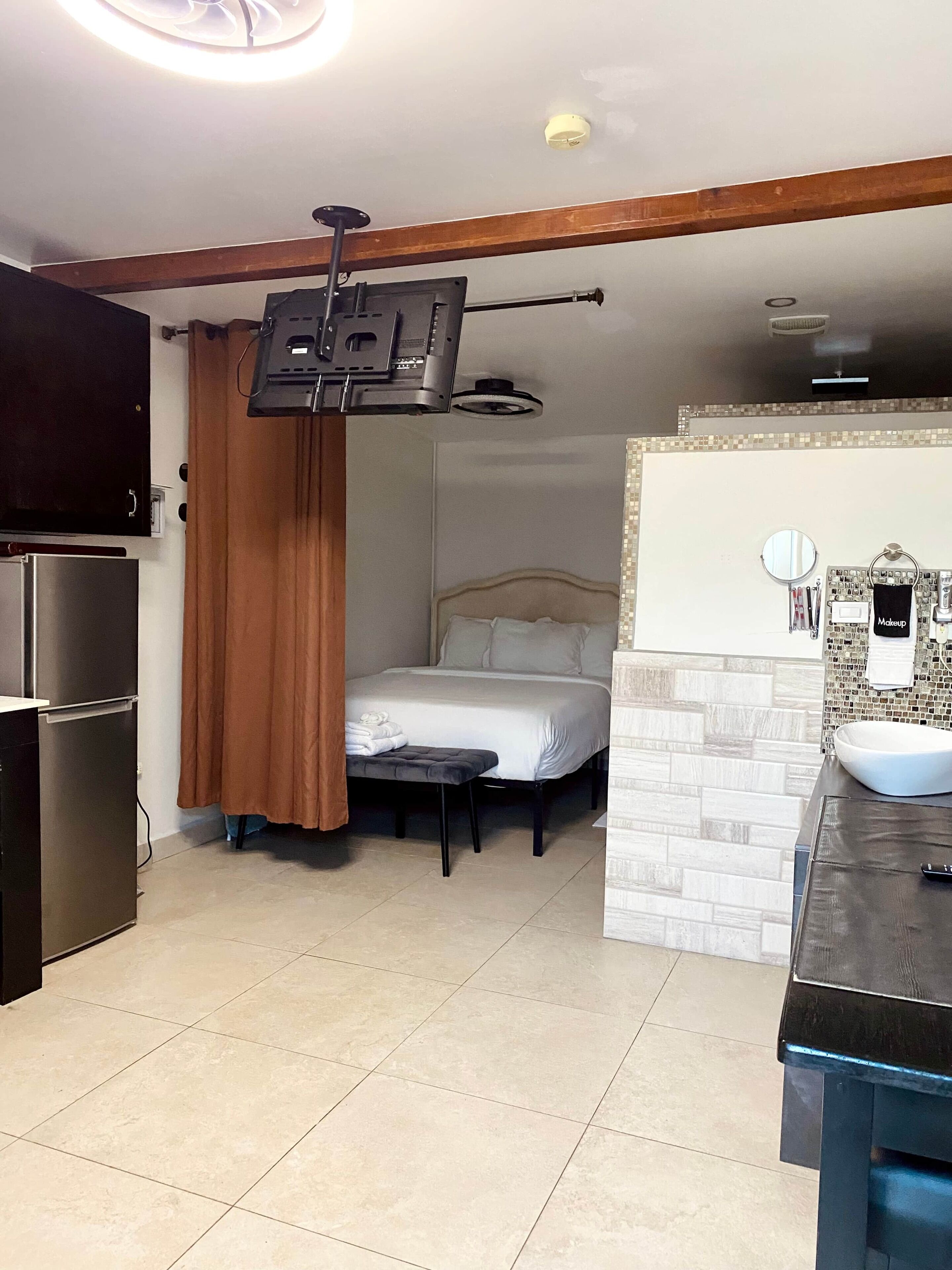 Una habitación pequeña con cama, una mesita, lavamanos integrado y televisor montado en la pared.