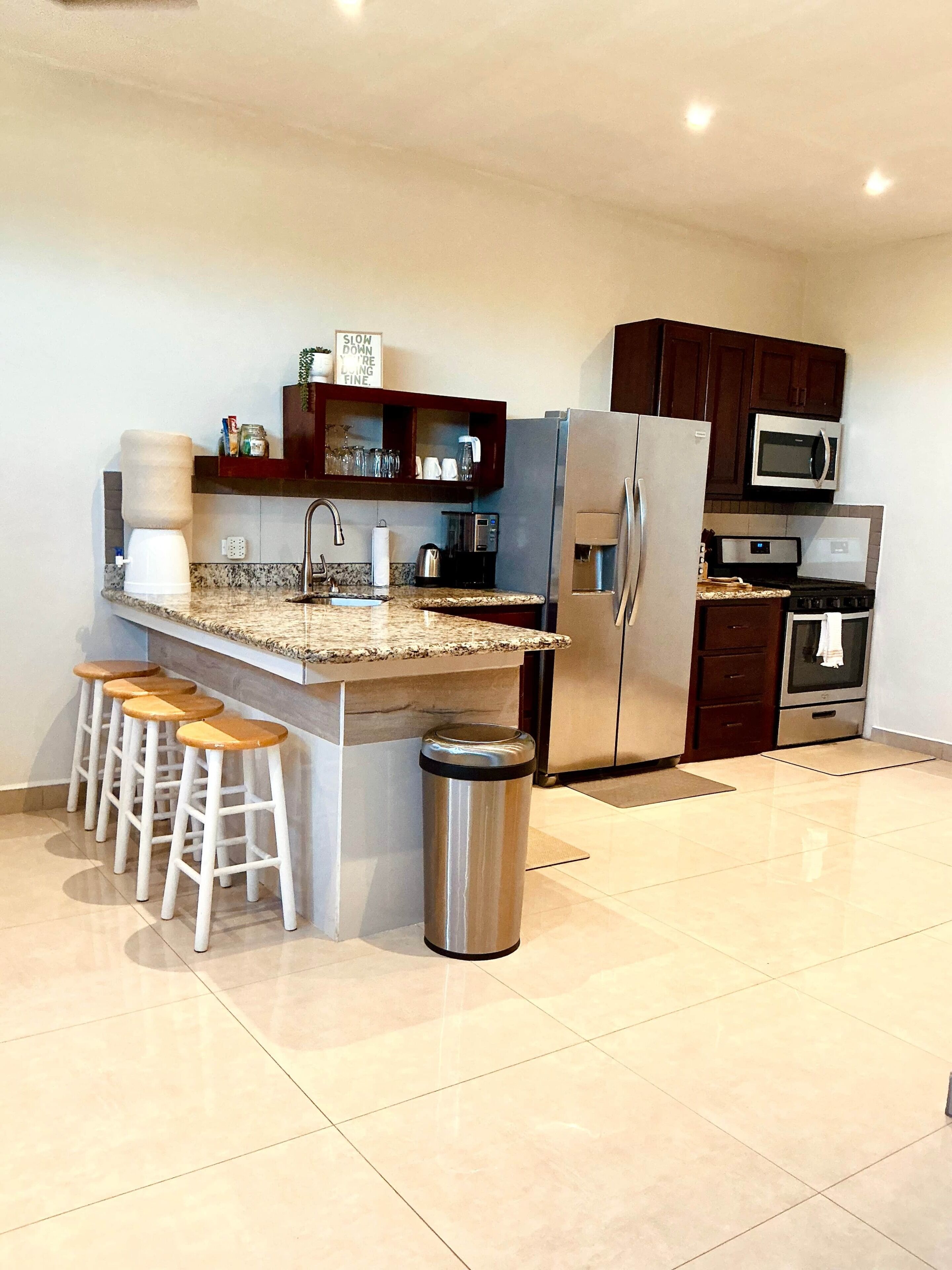 A modern kitchen with a central island, bar stools, a stainless steel trash can, and built-in appliances including a refrigerator, microwave, and oven.