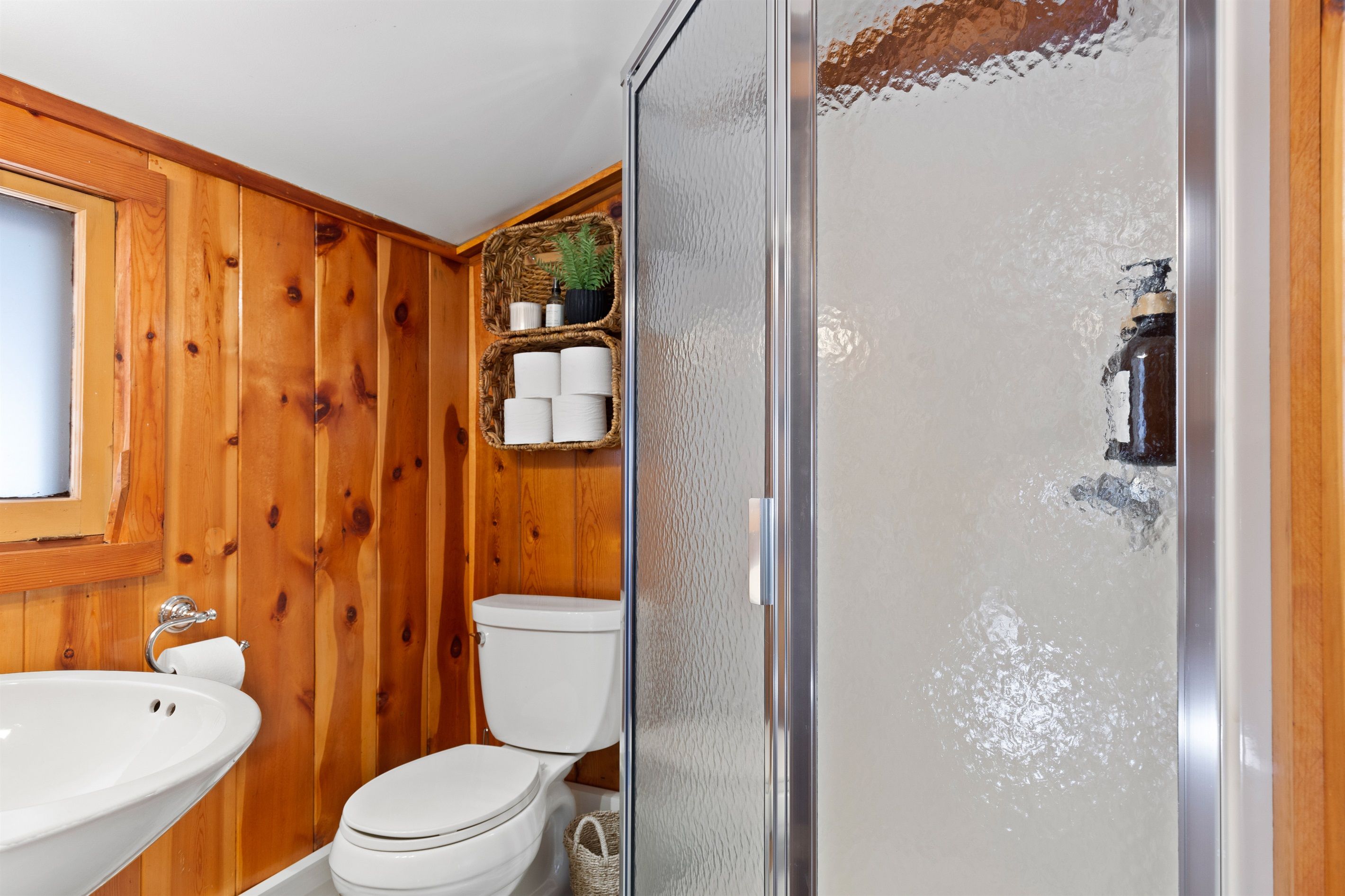 Un baño con paneles de madera, cabina de ducha de vidrio, lavabo blanco y un inodoro.