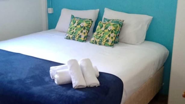 A neatly made bed with a blue blanket and white pillows, a framed picture on the wall, and a door to the left. - Legado das Aguas (Miracatu)
