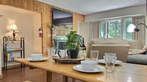 A cozy living room with a wooden dining table set for two, a sofa, and a television mounted on a wooden wall. - The Grizz- AC, Pool, Sauna, Gym, Near Resort (Killington)