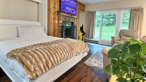 A bedroom with a large bed, a television mounted on the wall, a fireplace, and a view of the outdoors through large windows. - The Grizz- AC, Pool, Sauna, Gym, Near Resort (Killington)