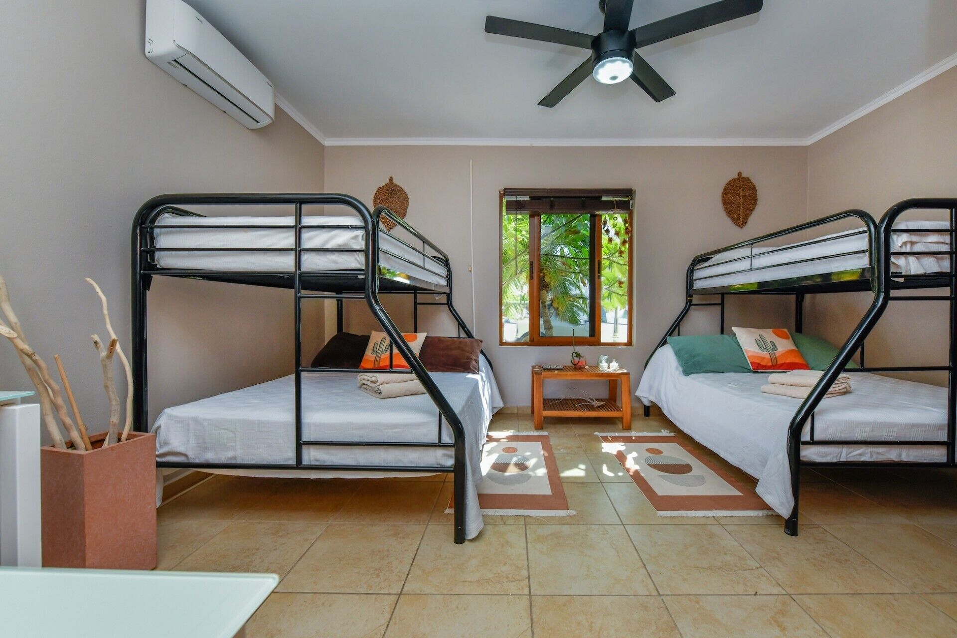 A room with two bunk beds, a ceiling fan, a window with a view of palm trees, and a small table with a plant.