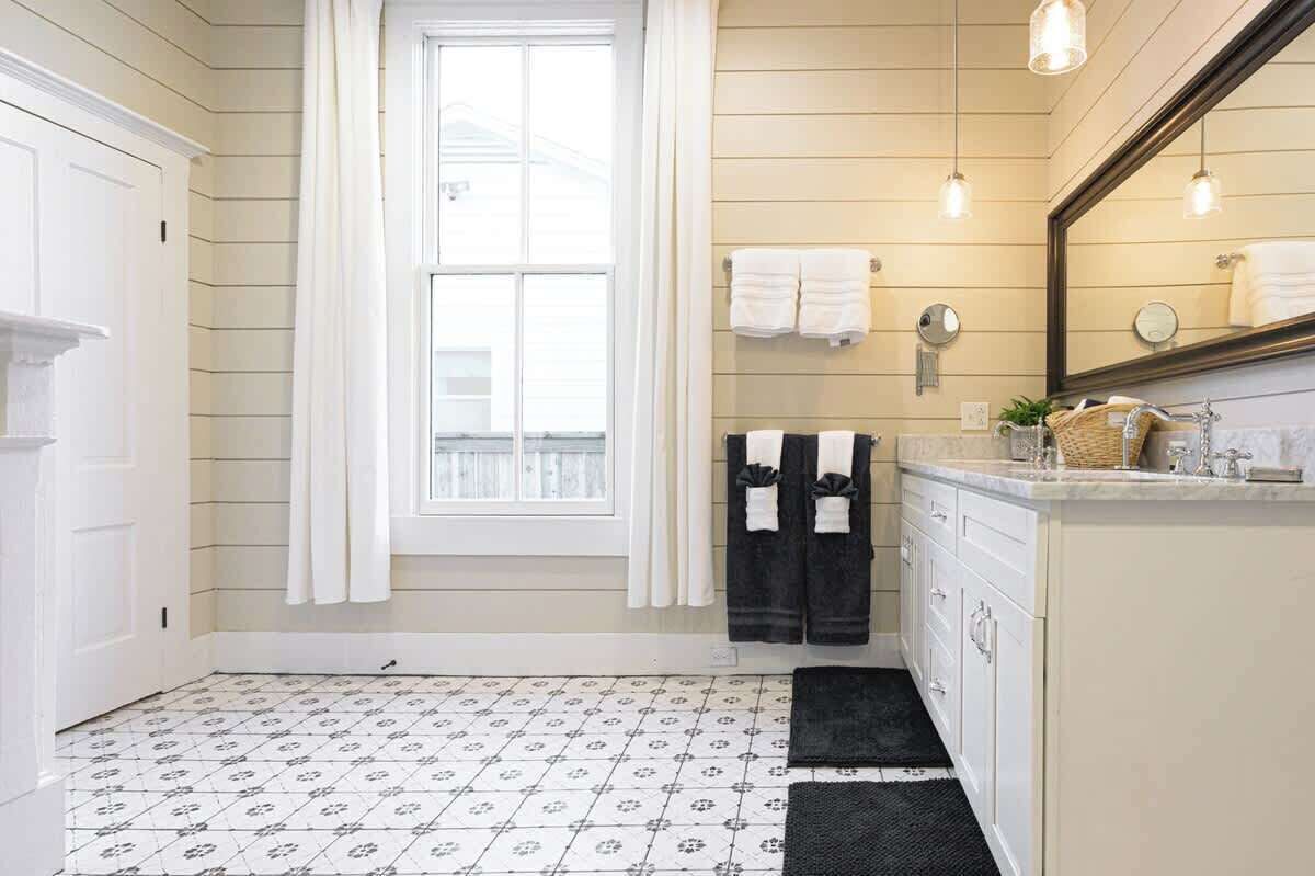 A bathroom with a window, white tiled floor, a sink with a mirror, and towels hanging on the wall.